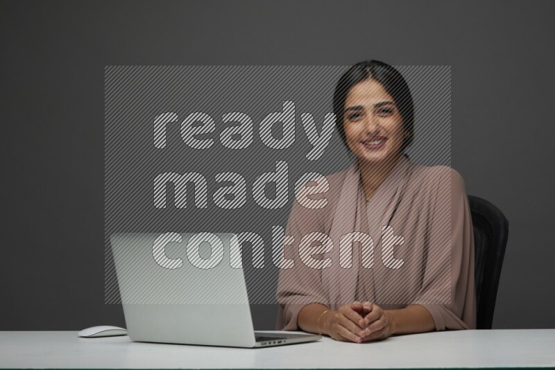 A Saudi woman Sitting on her desk on a Gray Background wearing Brown Abaya