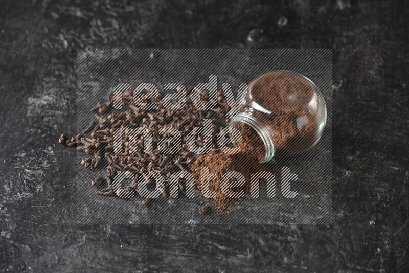 A flipped glass spice jar full of cloves powder and powder came out of it with cloves spread on textured black flooring