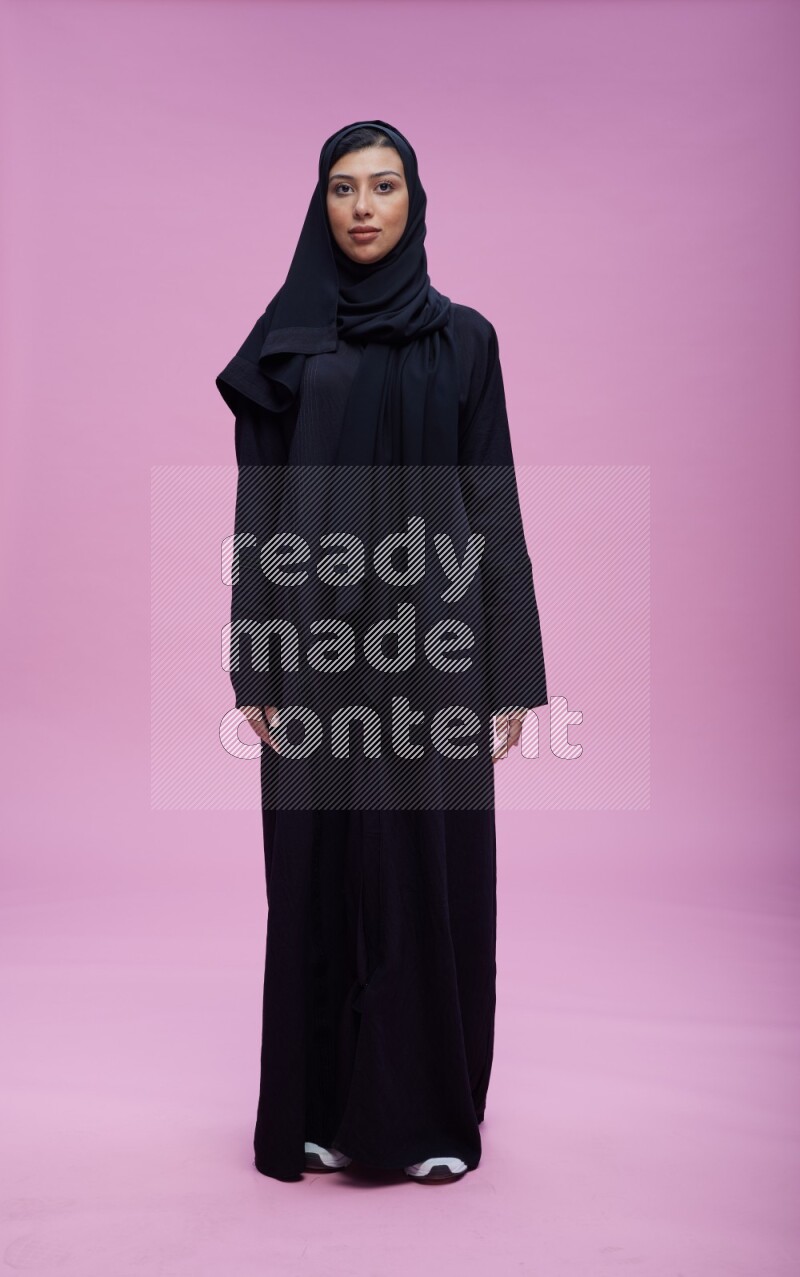 Saudi woman wearing Abaya standing interacting with the camera on pink background