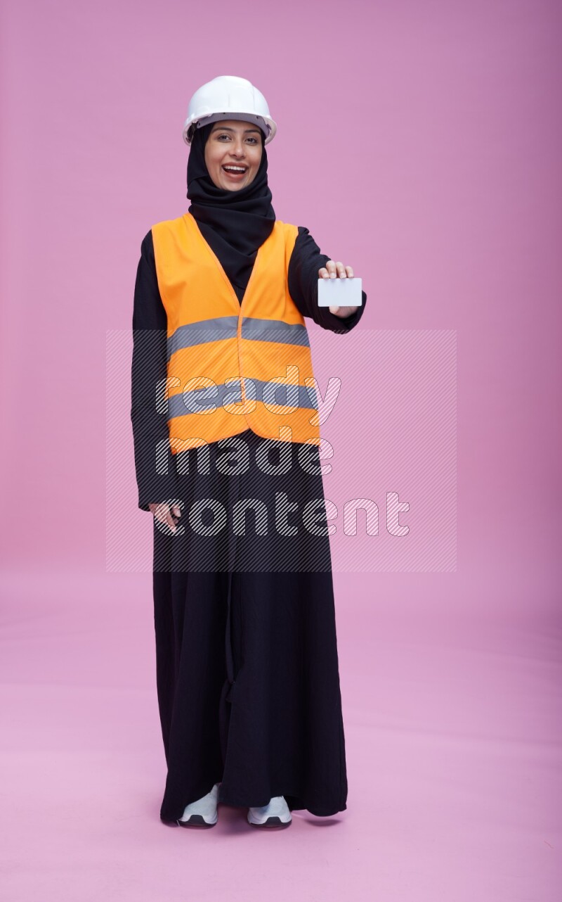 Saudi woman wearing Abaya with engineer vest and helmet standing holding ATM card on pink background