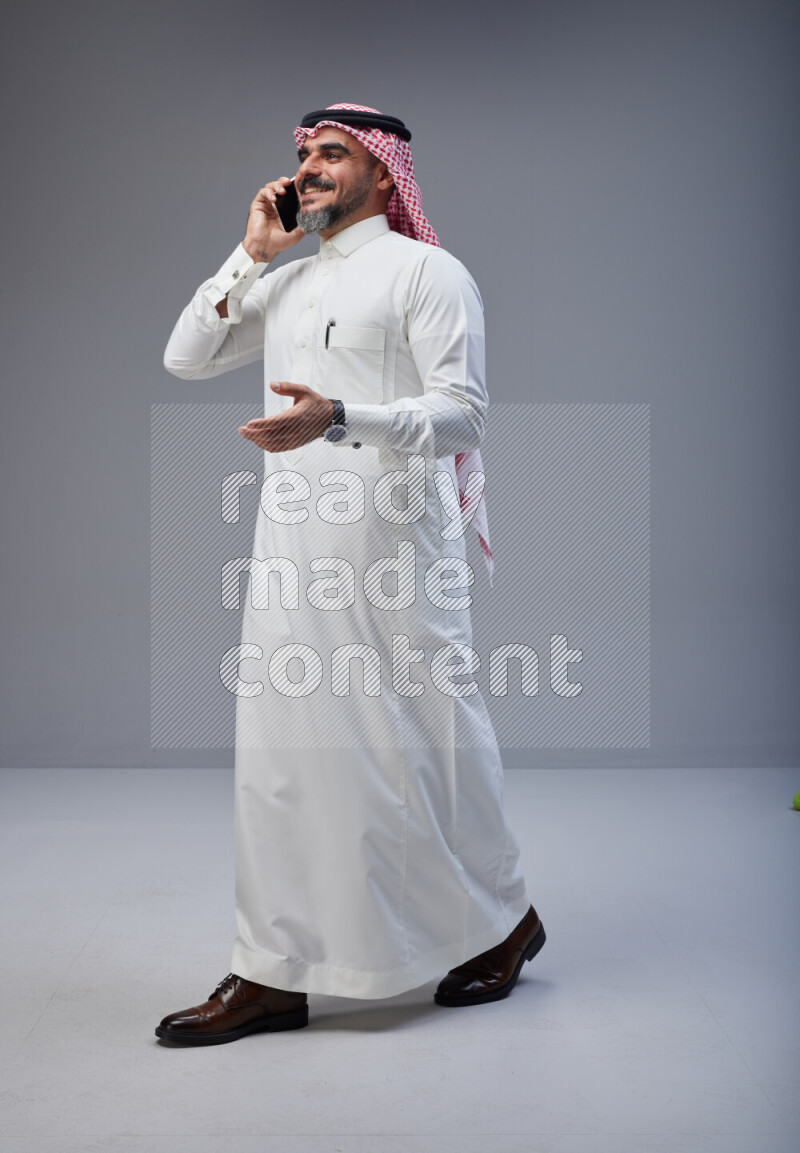 Saudi man Wearing Thob and red Shomag standing talking on phone on Gray background