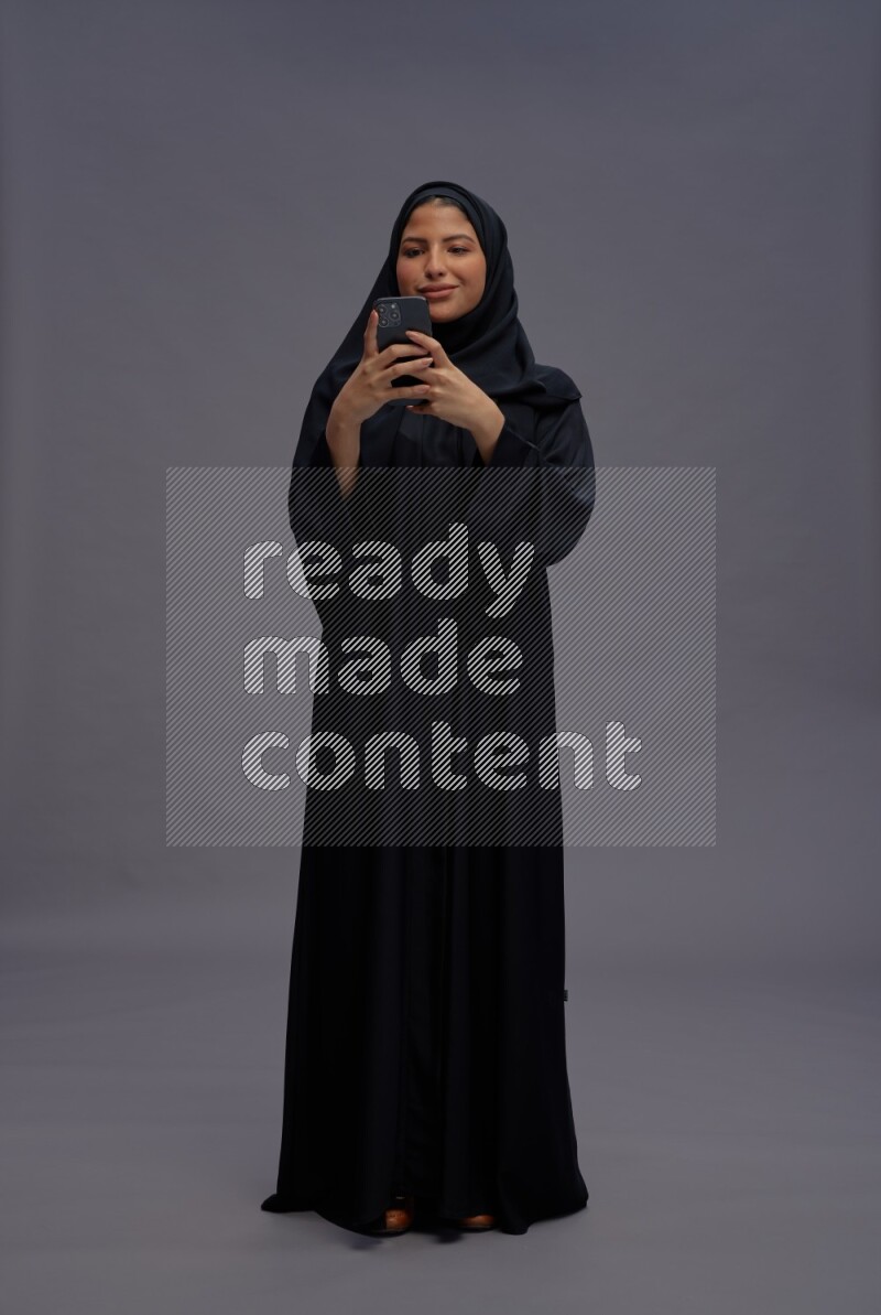 Saudi woman wearing Abaya standing texting on phone on gray background