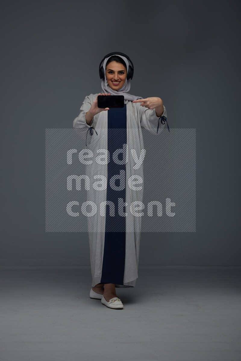 A Saudi woman wearing a light gray Abaya and head scarf setting on a dark grey chair and watching on the tablet eye level on a grey background