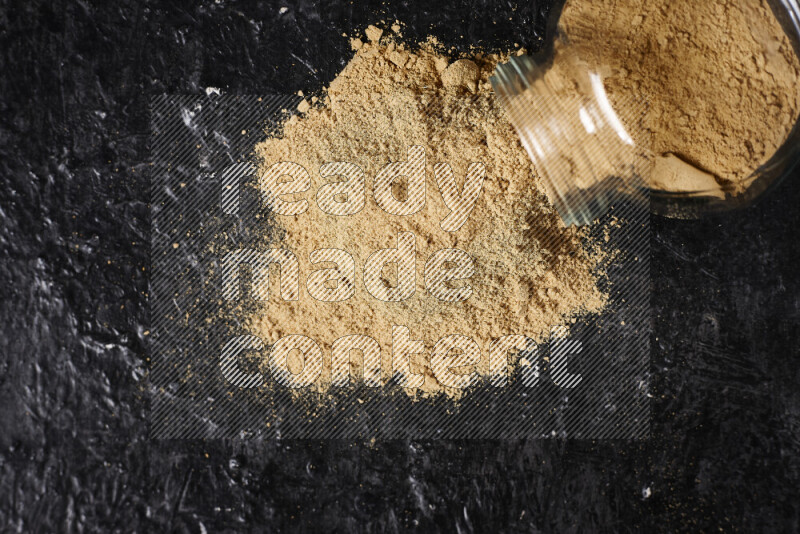 A glass jar full of ground ginger powder flipped with some spilling powder on black background