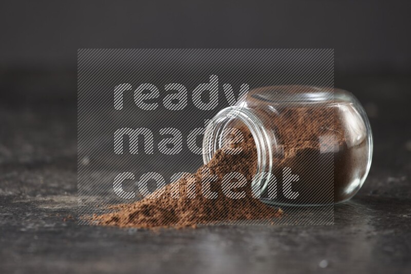 A flipped glass spice jar full of cloves powder and powder came out of it on textured black flooring