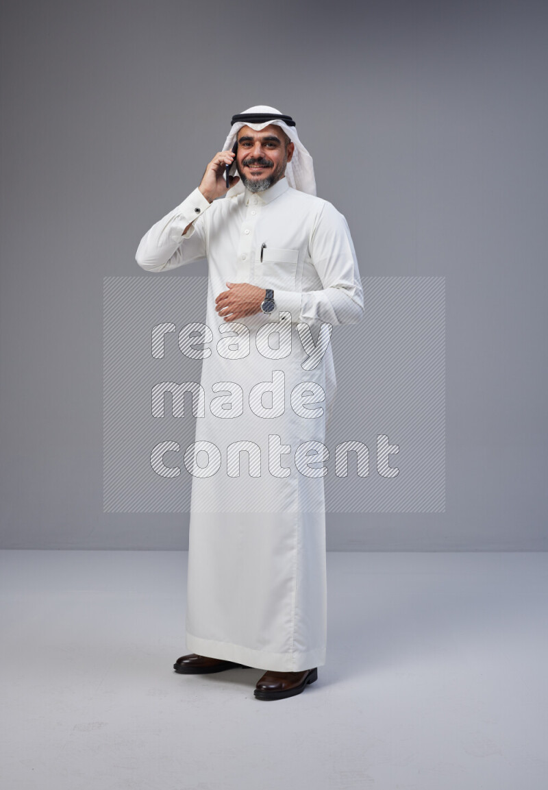 Saudi man Wearing Thob and white Shomag standing talking on phone on Gray background