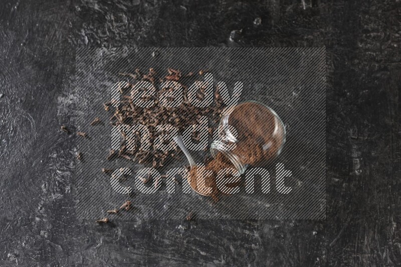 وعاء زجاجي للبهارات مقلوب وملعقة معدنية ممتلئان ببودرة القرنفل مع حبوب القرنفل المتناثرة على أرضية سوداء