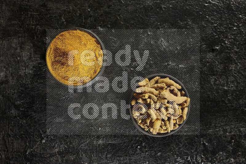 2 pottery black bowls, one full of turmeric powder and the other full of dried turmeric whole fingers on a textured black flooring