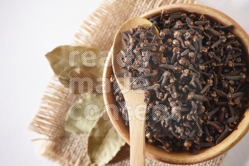 A wooden bowl and a wooden spoon full of cloves on a piece of burlap on a white flooring