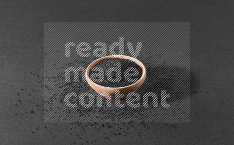 A wooden bowl full of black seeds surrounded by seeds on a black flooring