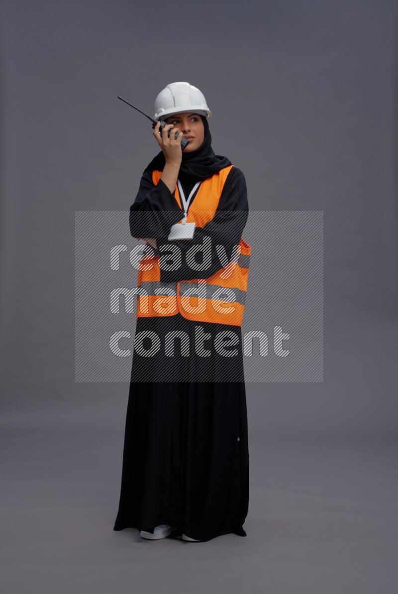 Saudi woman wearing Abaya with engineer vest with neck strap employee badge standing holding walkie-talkie on gray background