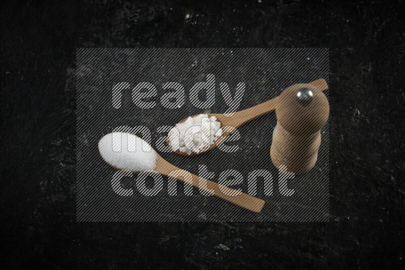 2 wooden spoons filled with fine and coarse salt with wooden grinder beside them on black background