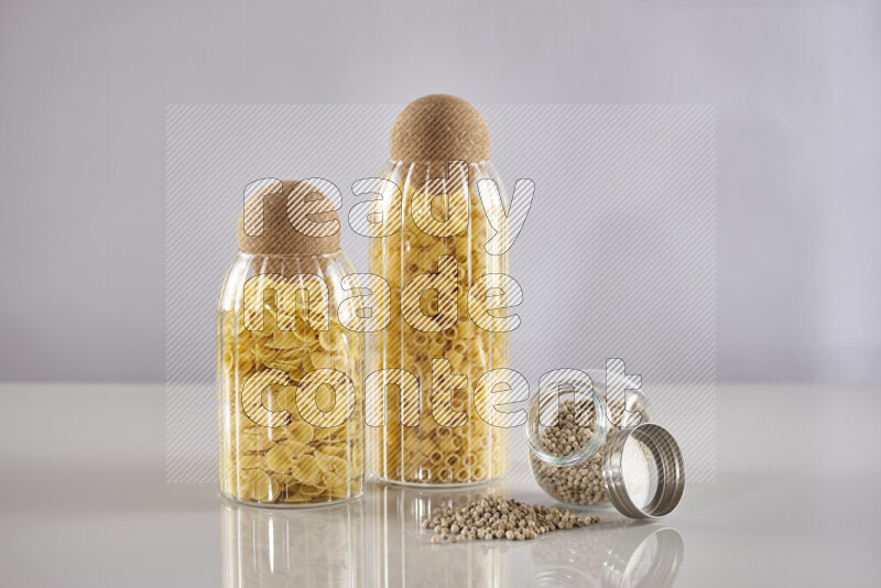 Raw pasta in glass jars with white peppers on light grey background