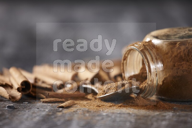 Herbal glass jar full cinnamon powder flipped and a metal spoon full of powder surrounded by cinnamon sticks on textured black background in different angles