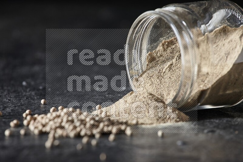 A flipped glass jar full of white pepper powder with spilled powder and pepper beads on textured black flooring