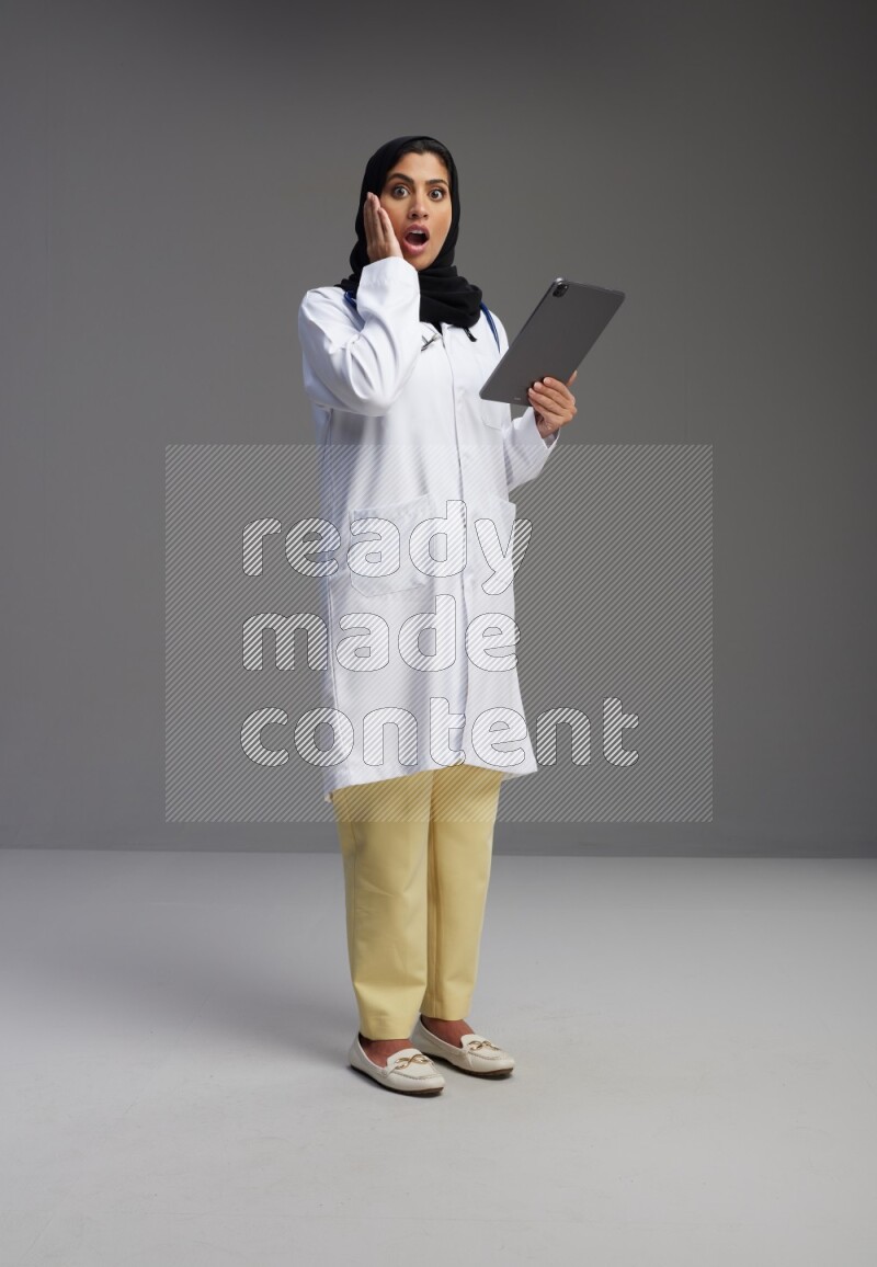 Saudi woman wearing lab coat with stethoscope standing working on tablet on Gray background