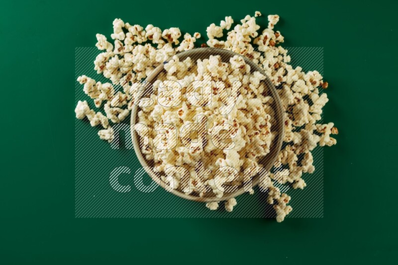 A beige ceramic bowl full of popcorn with popcorn beside it on a green background in a top view shot