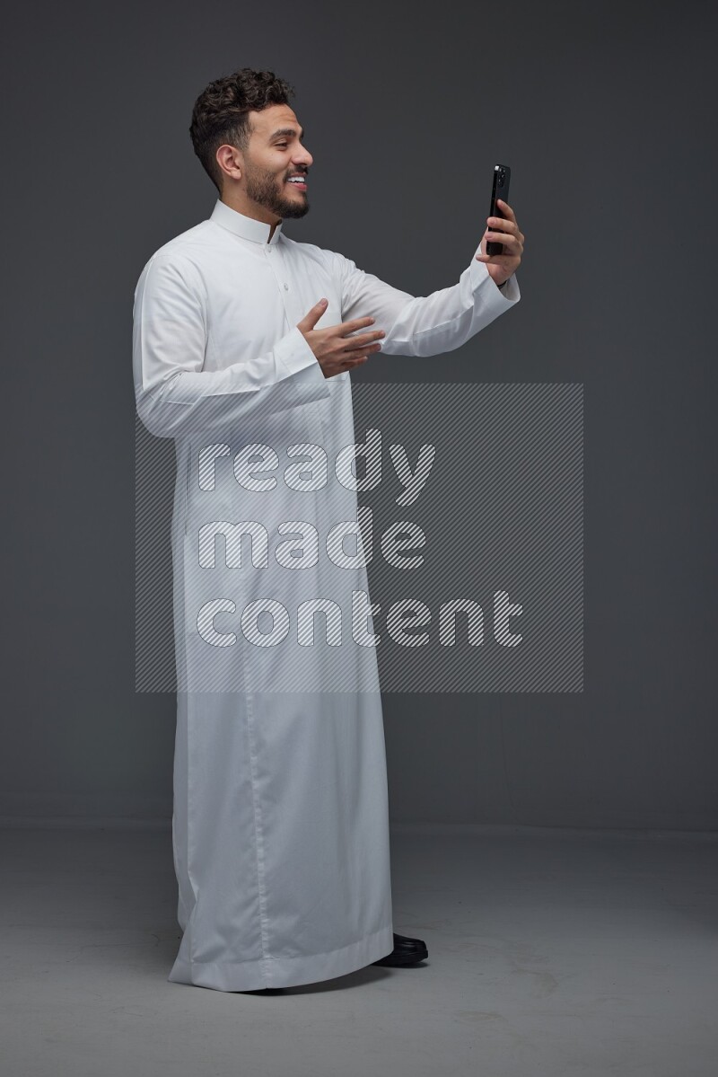 A Saudi man wearing Thobe and taking selfie with his phone making different poses eye level on a gray background