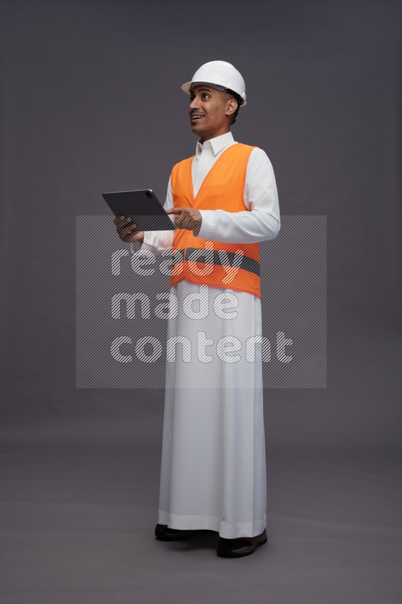 Saudi man wearing thob with engineer vest standing working on tablet on gray background