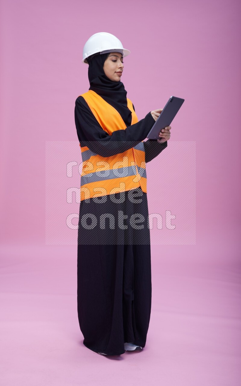 Saudi woman wearing Abaya with engineer vest and helmet standing working on tablet on pink background