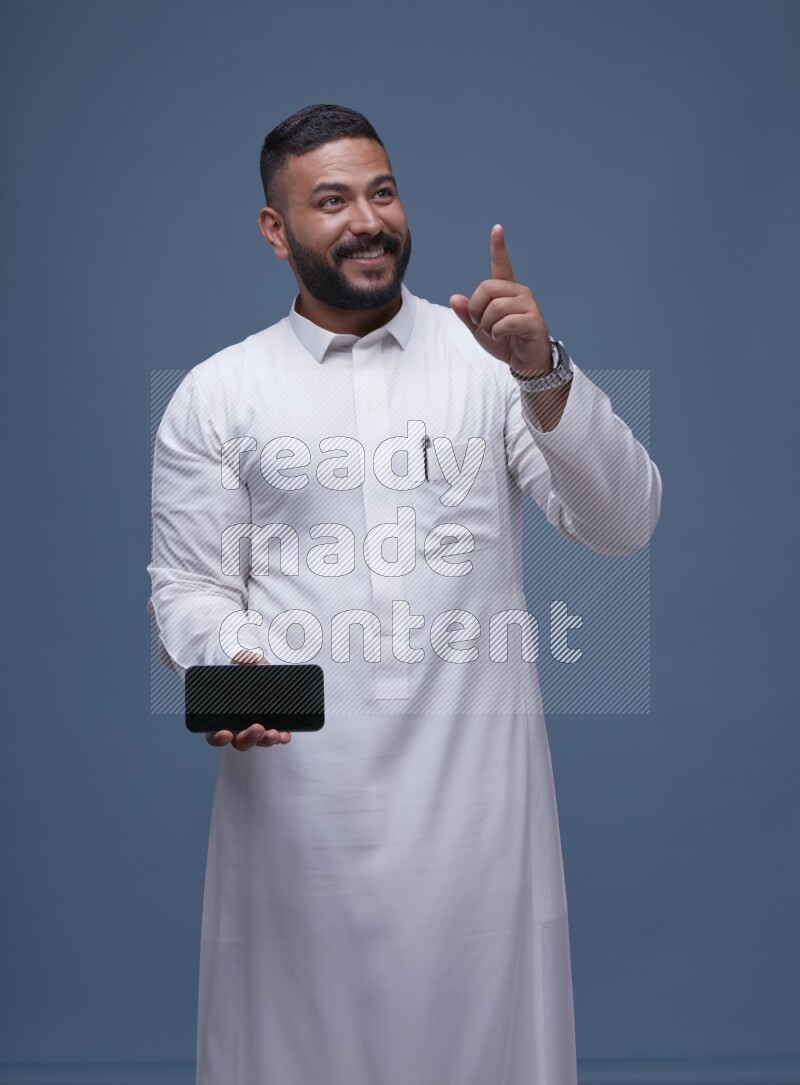 A man Showing his phone in Blue Background wearing Saudi Thob