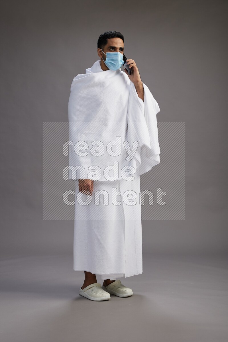 A man wearing Ehram with face mask Standing talking on phone on gray background