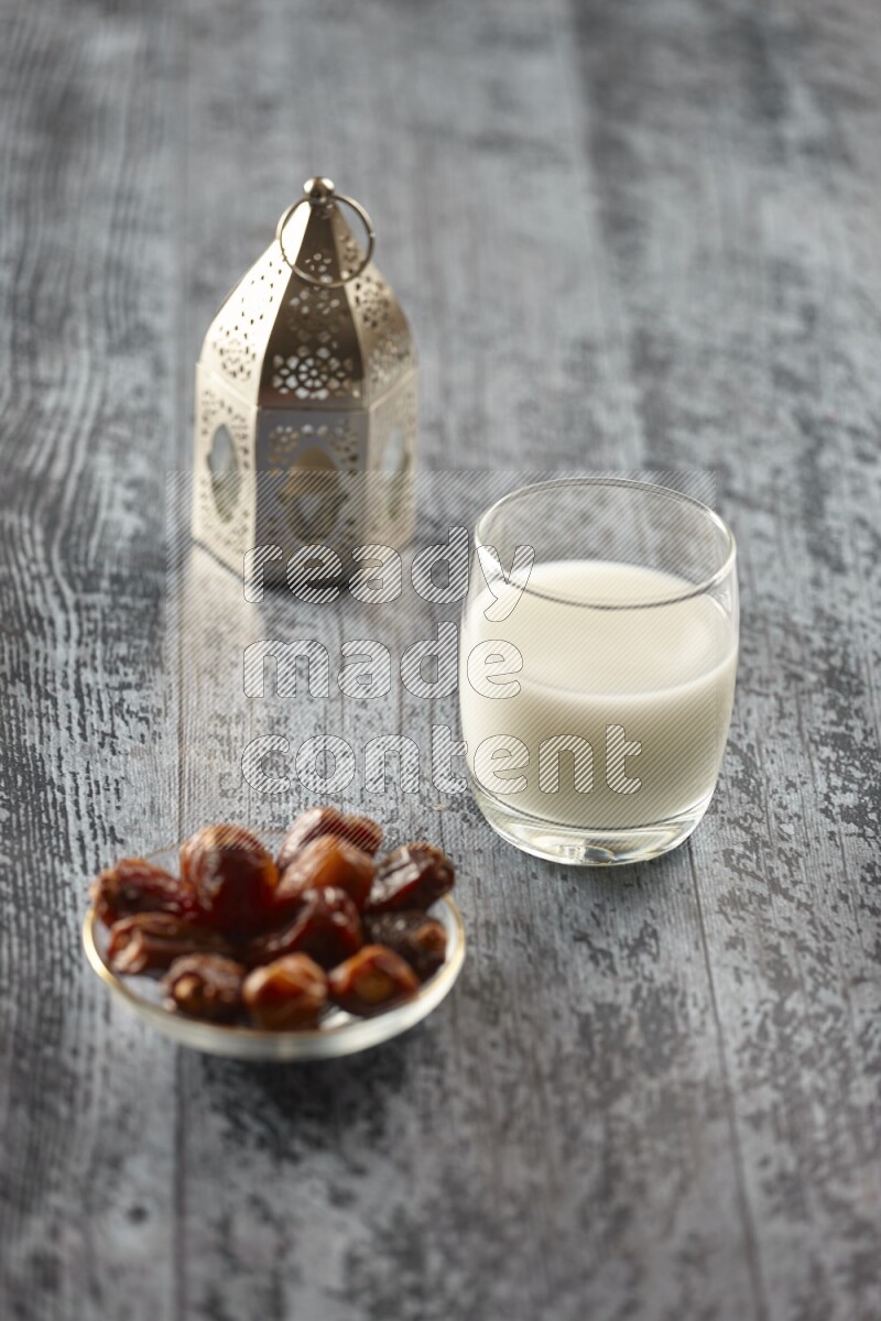 A silver lantern with different drinks, dates, nuts, prayer beads and quran on grey wooden background