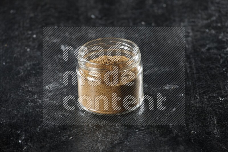 A glass jar full of cumin powder on a textured black flooring