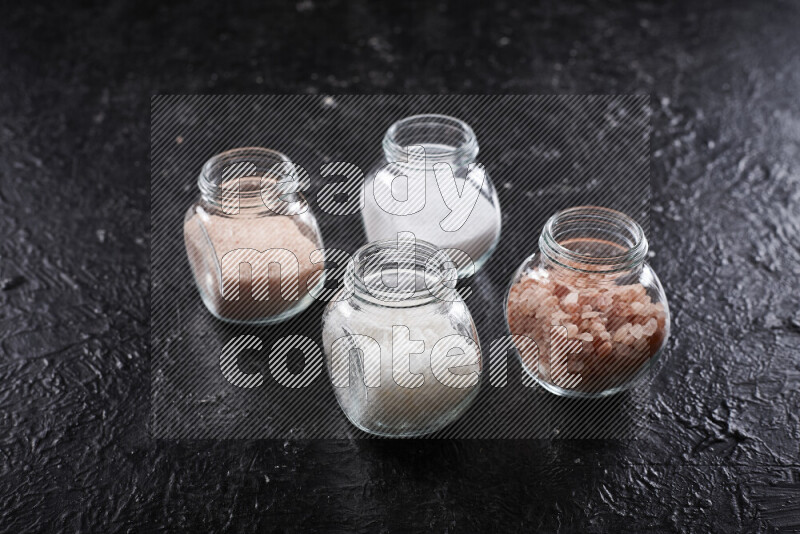 4 glass jars filled with table salt, coarse sea salt, fine himalayan salt and coarse himalayan salt on black background