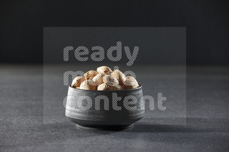 A black pottery bowl full of almonds on a black background in different angles