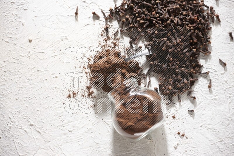 A flipped glass spice jar and a metal spoon full of cloves powder and powder came out of the jar with cloves spread on textured white flooring