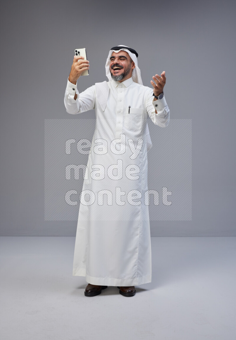 Saudi man Wearing Thob and white Shomag standing texting on phone on Gray background
