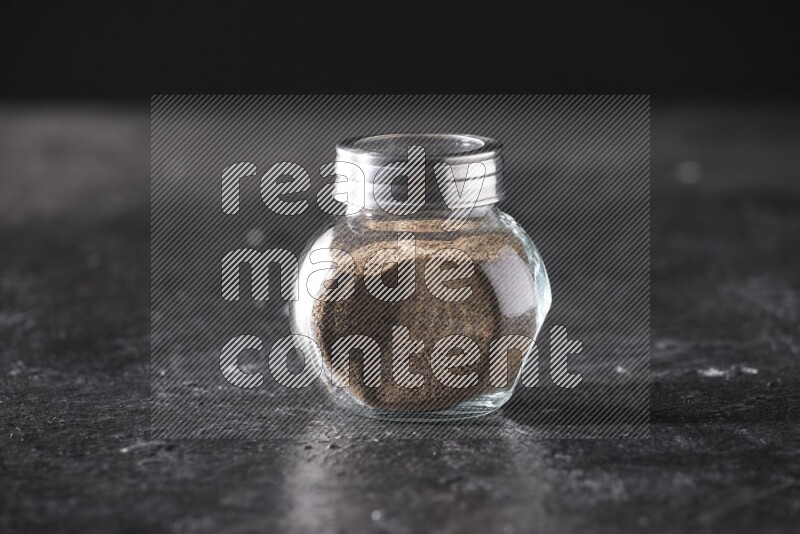A glass spice jar full of black pepper powder on textured black flooring
