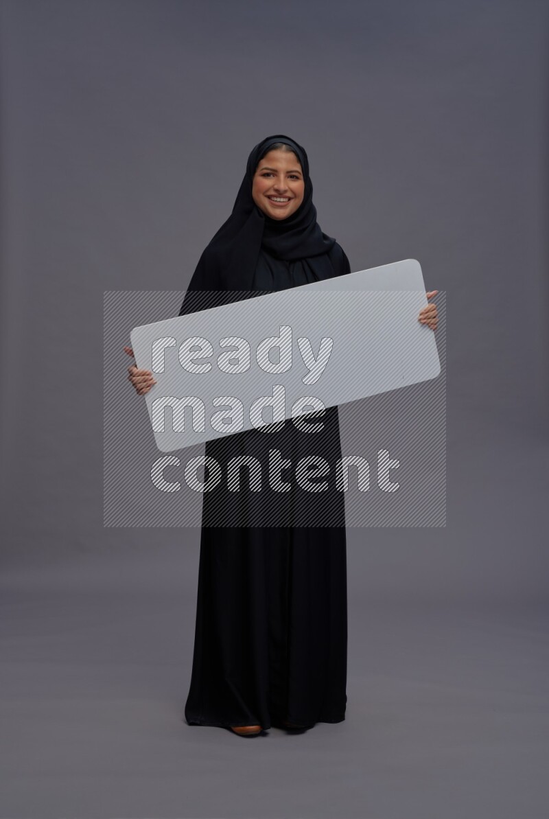 Saudi woman wearing Abaya standing holding white board on gray background