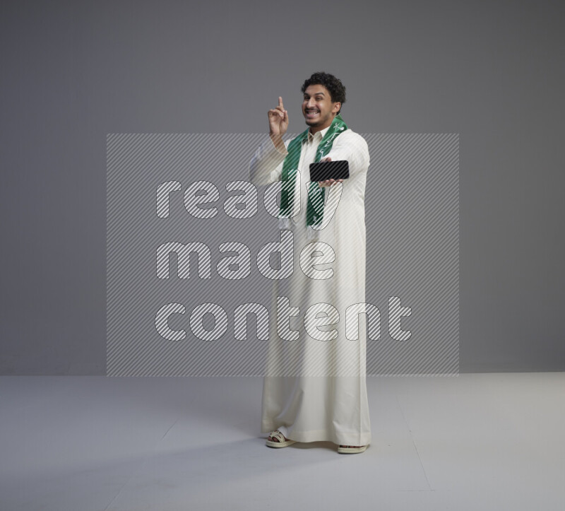 A Saudi man standing wearing thob and Saudi flag scarf showing phone to camera on gray background