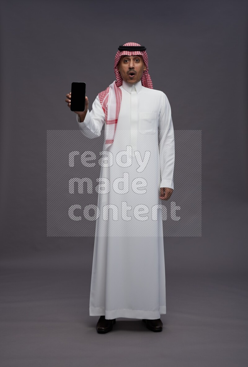 Saudi man wearing thob and shomag standing showing phone to camera on gray background