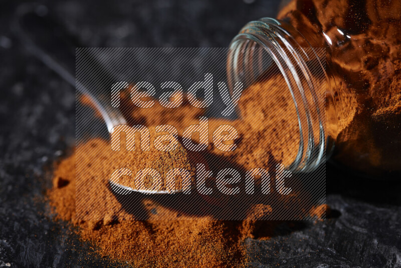 A glass jar full of ground paprika powder flipped with some spilling powder on black background
