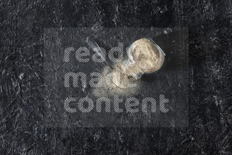 A flipped herbal glass jar and a metal spoon full of white pepper powder with spilled powder on textured black flooring