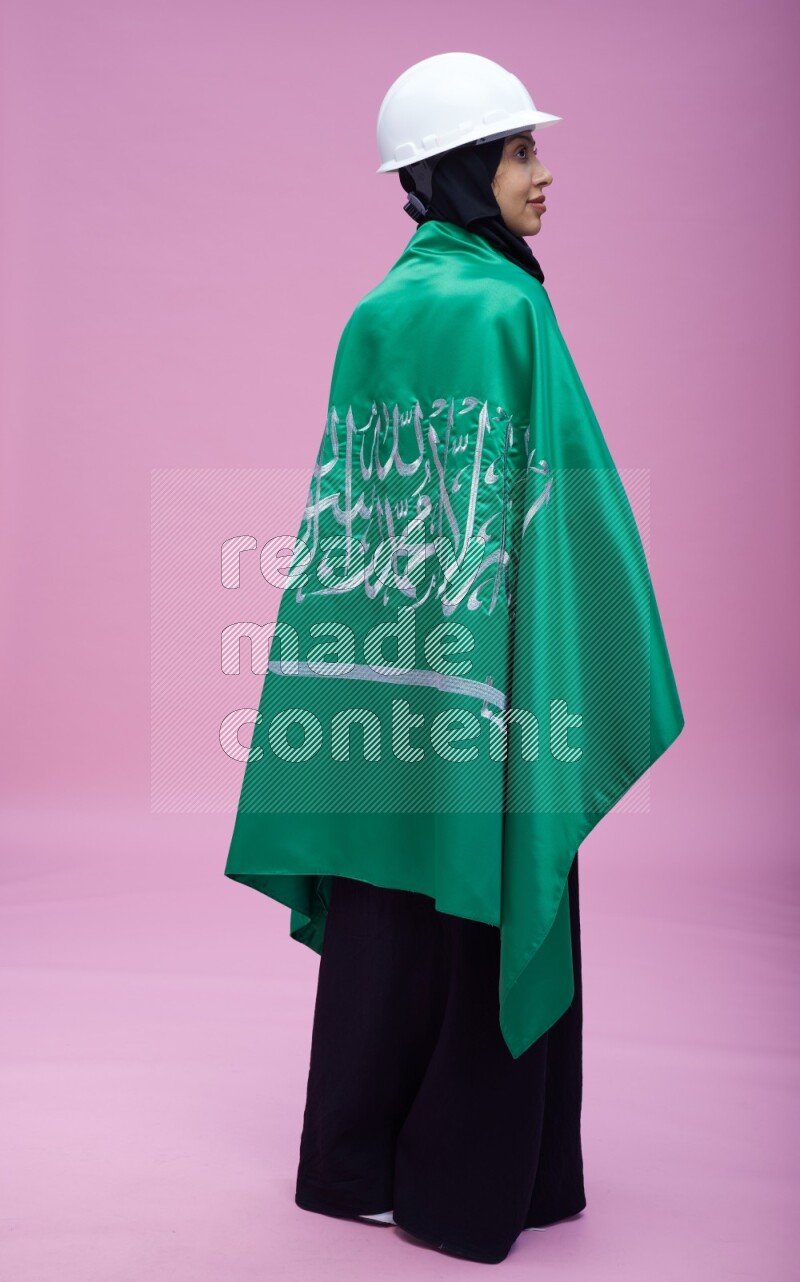 Saudi woman wearing Abaya with engineer vest and helmet standing holding Saudi flag on pink background
