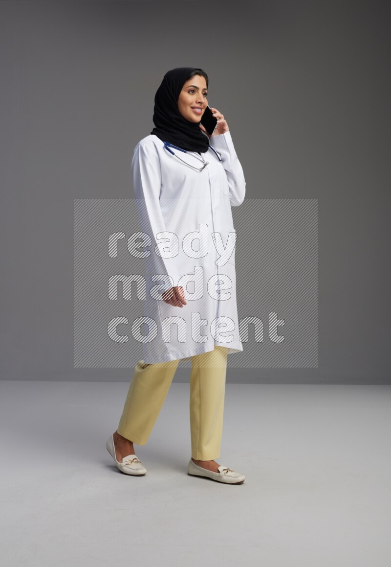 Saudi woman wearing lab coat with stethoscope standing talking on phone on Gray background