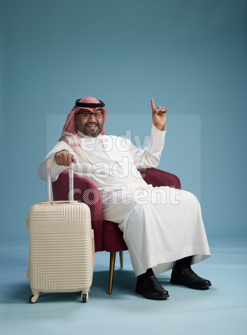 Saudi Man with shimag sitting on chair with a travel luggage on blue background