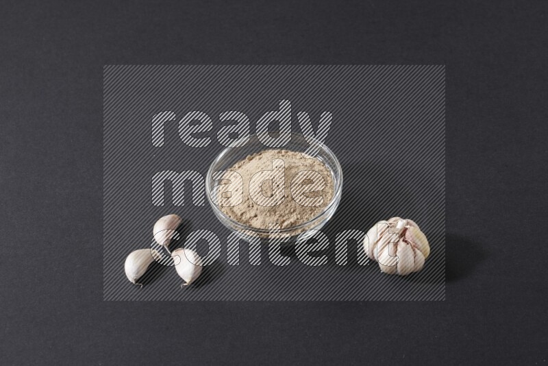 وعاء زجاجي ممتلئ ببودرة الثوم مع بعض فصوص الثوم ورأس ثوم كاملة على خلفية سوداء