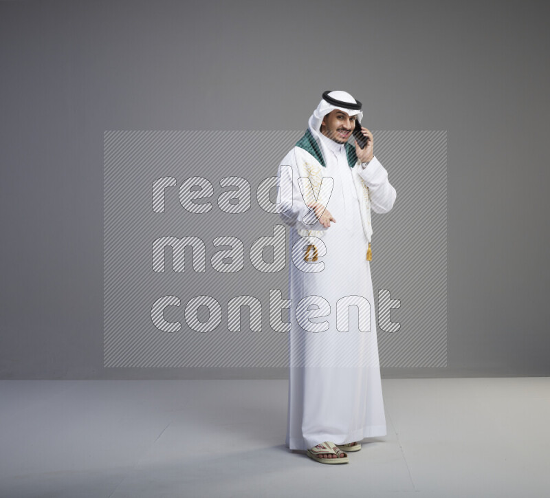 A saudi man standing wearing thob and white shomag with flag scarf on his neck talking on phone on gray background