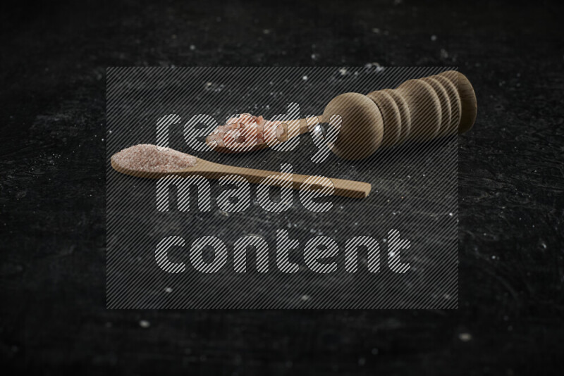 2 wooden spoons filled with fine and coarse salt with wooden grinder beside them on black background