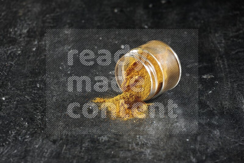 A flipped glass jar full of turmeric powder and powder spilled out of it on a textured black flooring