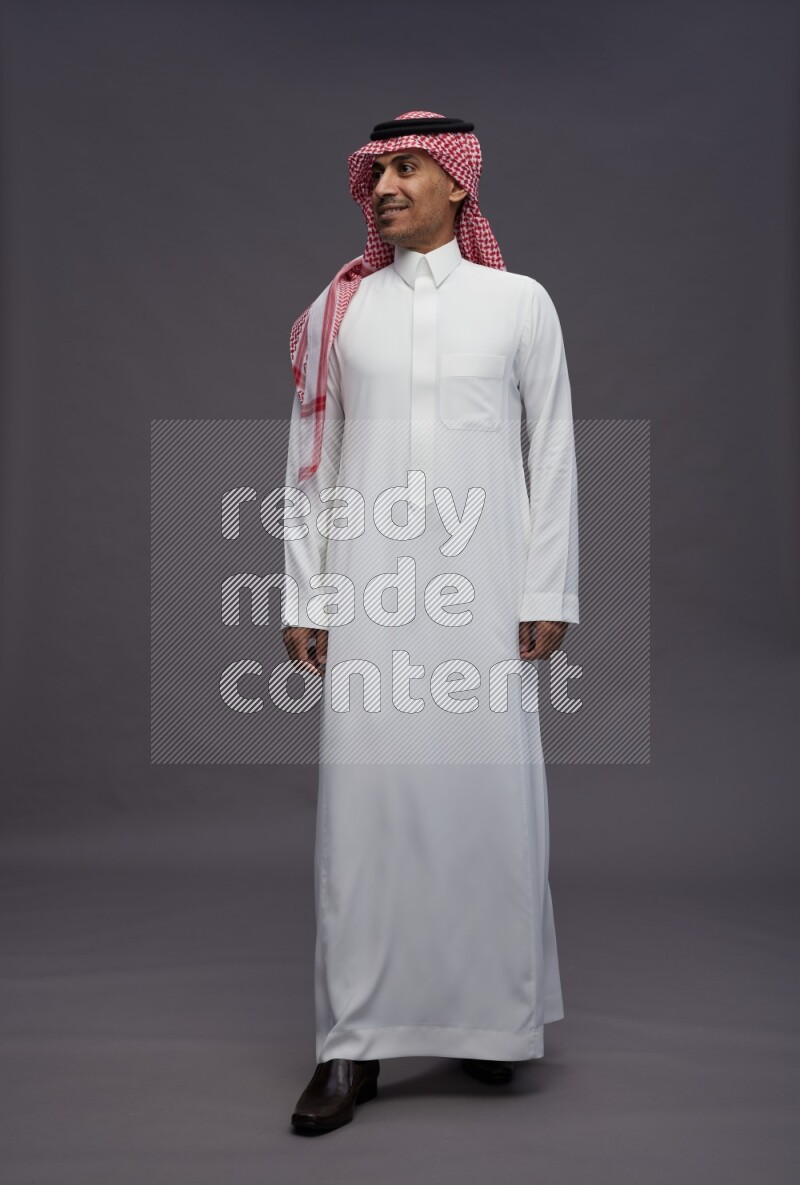 Saudi man wearing thob and shomag standing interacting with the camera on gray background