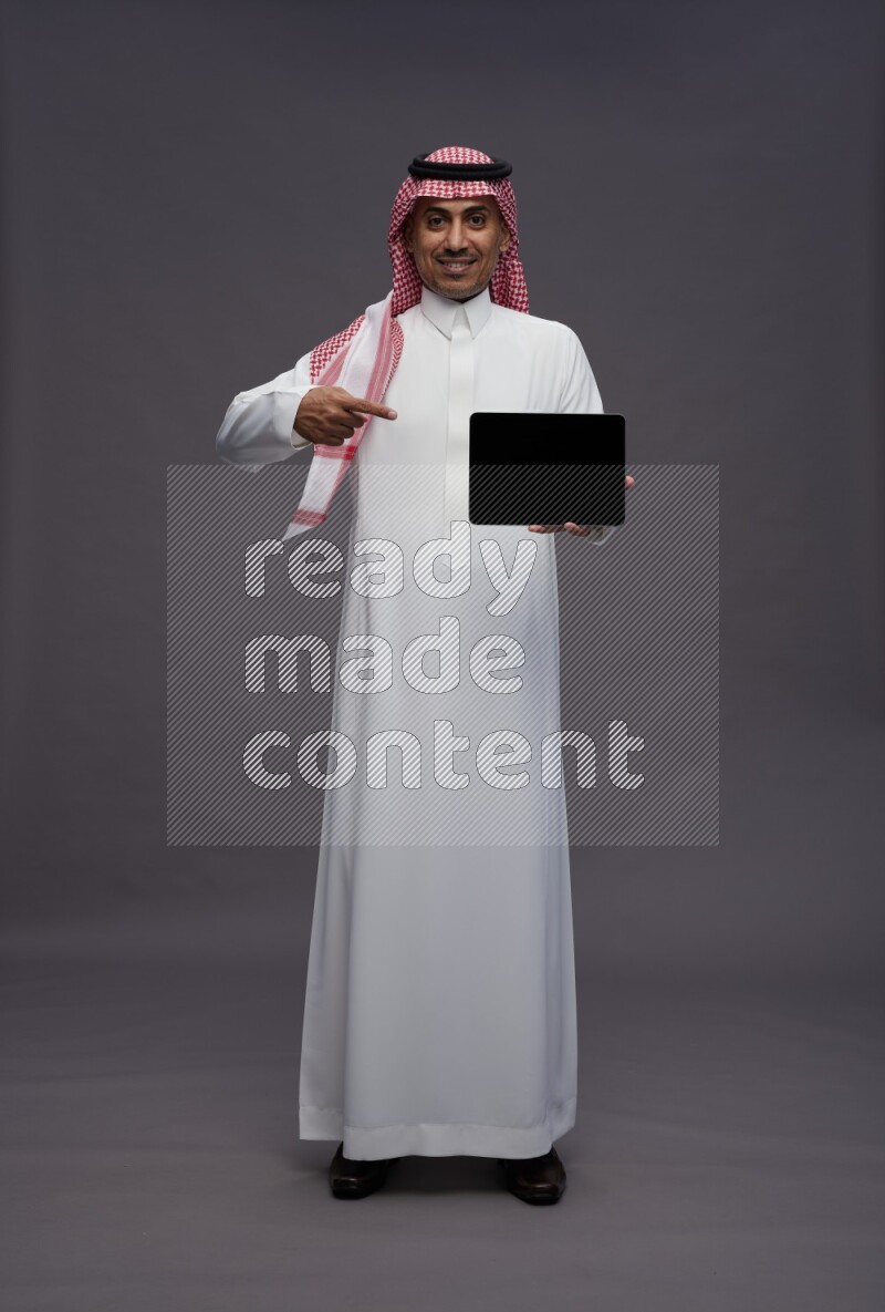 Saudi man wearing thob and shomag standing showing tablet to camera on gray background