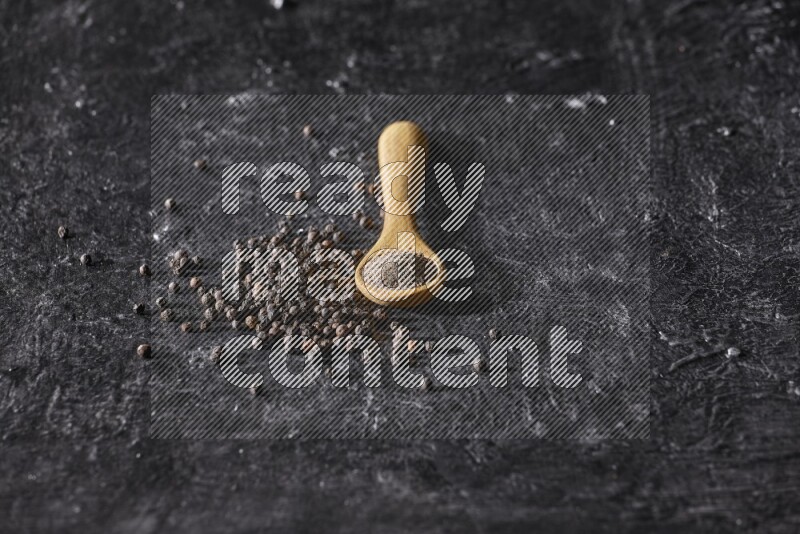 A wooden spoon full of black pepper powder and black pepper beads spread on a textured black flooring