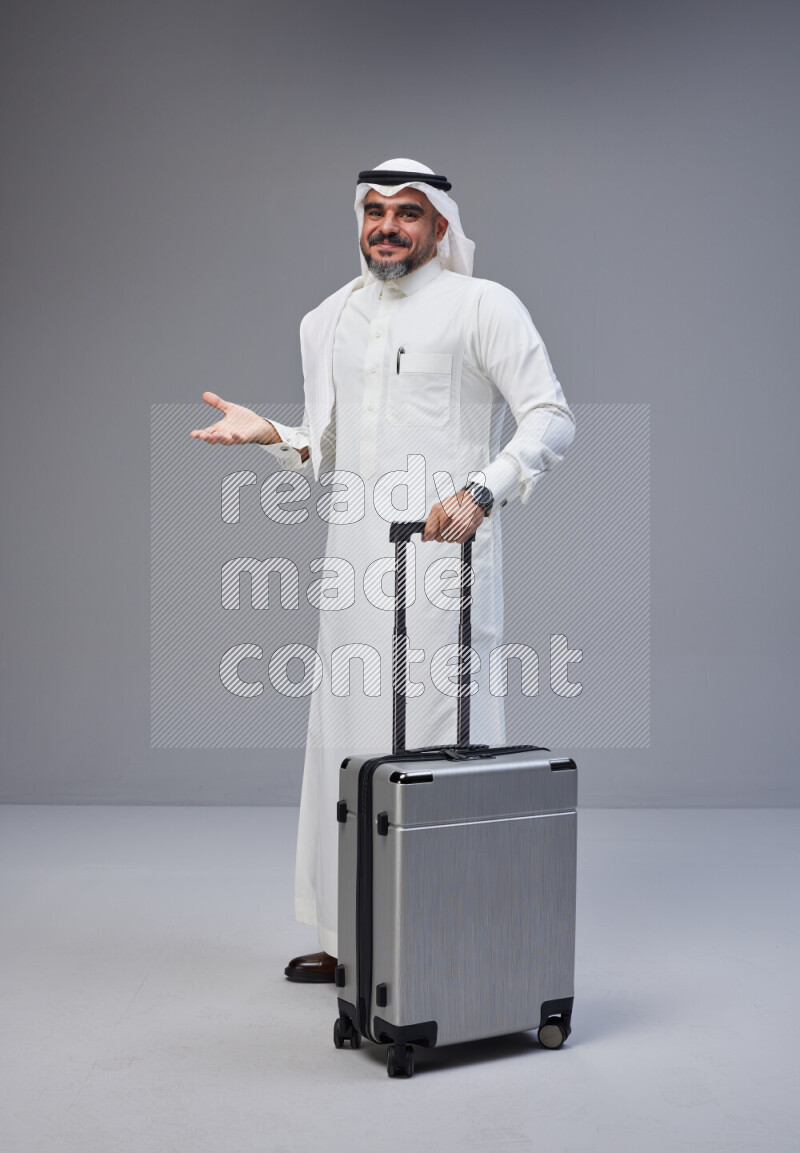 Saudi man wearing Thob and white Shomag standing holding Travel bag on Gray background