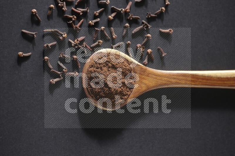 A wooden ladle full of cloves powder and some of whole cloves around it on a black flooring